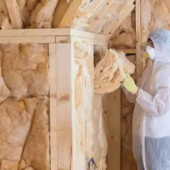 Ocieplenie domu drewnianego od zewnątrz: pracownik w kombinezonie montuje wełnę mineralną w drewnianej konstrukcji.