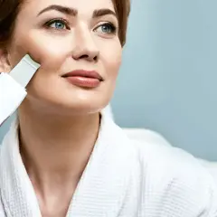 Kobieta w białym szlafroku wykonuje peeling kawitacyjny w domu, poprawiając wygląd swojej skóry.