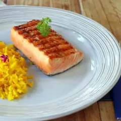 Un delicioso salmón a la plancha con marcas de parrilla, servido con arroz amarillo y decorado con perejil y flores moradas.