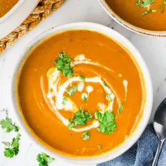A creamy, vibrant bowl of kabocha squash soup, swirled with coconut cream and garnished with fresh cilantro. A spoon rests on a blue napkin beside it.