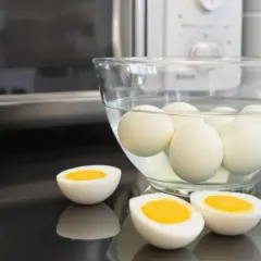 Des œufs durs dans un bol d'eau, prêts à être dégustés. Un œuf est coupé en deux, révélant un jaune d'or parfait. La méthode de cuisson par oeuf dur micro onde est rapide et facile.