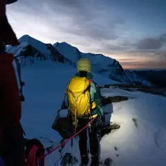 Alpinista z żółtym plecakiem lawinowym, gotowy na wyprawę w góry o wschodzie słońca. Plecak lawinowy jak działa? Zapewnia bezpieczeństwo w górach.