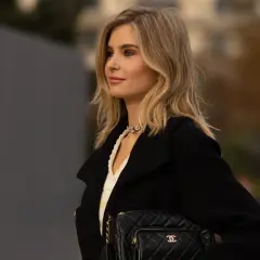 Kobieta z beżowym blondem, w czarnym płaszczu i z pikowaną torebką Chanel, prezentuje elegancką stylizację.