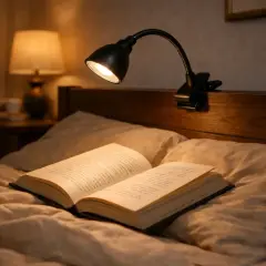 Un libro abierto descansa sobre almohadas, iluminado por un foco pinza negro.