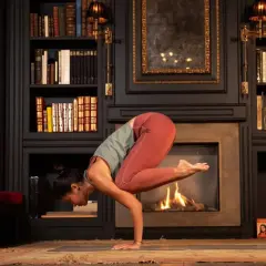 Mujer en postura de grulla, inspirada en frases yoga Patanjali. Chimenea encendida y estanterías llenas de libros.