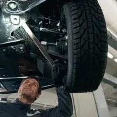 Mechanik bada podwozie samochodu, szukając przyczyny stukania w kole. Używa latarki, by dokładnie obejrzeć elementy zawieszenia.