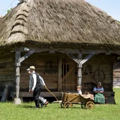 Drewniana chata z krytym strzechą dachem, mężczyzna ciągnie wóz z dzieckiem. Atrakcje dla dorosłych w skansenie.