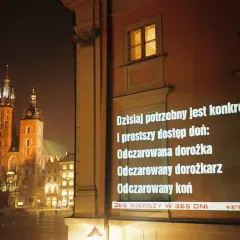 Nocny Kraków. Na ścianie budynku wyświetlane są wiersze o krakowie, m.in. o odczarowanej dorożce.