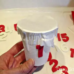 Un bicchiere decorato con il numero 1, pronto per essere parte di un calendario dell'avvento fai da te. Numeri sparsi sullo sfondo.