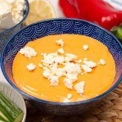 Cremiger Paprika-Feta-Dip in blauer Schüssel, garniert mit Feta-Käse. Dazu Gurkensticks und rote Paprika.