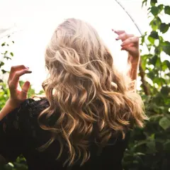 Blond włosy falują na wietrze, jakby przechodziły anwen test na porowatość. Kobieta w czarnej koronkowej bluzce stoi wśród zielonych liści.