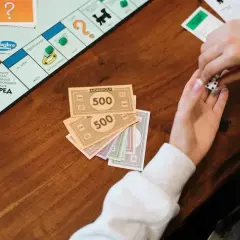 Ręce rzucają kostką do gry w Monopoly. Na stole leżą pieniądze z gry, symbolizujące eurobiznes i bogactwo.