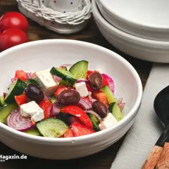 Frischer griechischer Salat mit Tomaten, Gurken, roten Zwiebeln, Oliven und Feta in einer weißen Schüssel.