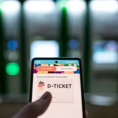Smartphone zeigt "D-TICKET" an, bereit für das Deutschlandticket per Lastschrift. Im Hintergrund unscharfe Automaten.