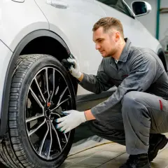 Mécanicien en combinaison grise, avec des gants, examine la roue d'une voiture blanche. Il semble chercher comment dégripper un étrier de frein sans démonter.