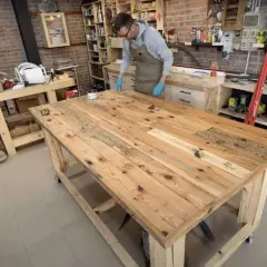 Uomo con guanti blu applica finitura su un robusto banco da lavoro in legno fai da te, circondato da attrezzi e materiali in un laboratorio.