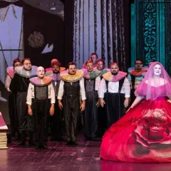 Aktorzy w barwnych kołnierzach i kobieta w sukni z różą na scenie operowej.