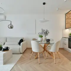 Nowoczesna aranżacja salonu z wygodną sofą, okrągłym stołem i aneksem kuchennym. Jasne kolory i drewniana podłoga tworzą przytulną atmosferę.