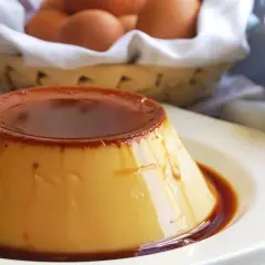 Delicioso flan de huevo casero con caramelo líquido, decorado con menta fresca. En el fondo, una cesta con huevos y un frasco de mermelada.