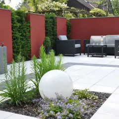 Moderner Garten ohne Rasen mit roten Mauern, Sitzgruppe und weißer Kugel im Blumenbeet.