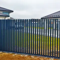 Nowoczesne ogrodzenia grzebieniowe w ciemnym kolorze, chroniące posesję z domami w tle.