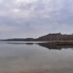 Spokojne jezioro Kamionki odbija pochmurne niebo i lasy. Drewniane molo wchodzi w głąb wody.