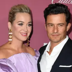 Katy Perry i Orlando Bloom na tle różowej ściany. Ona w fioletowej sukni, on w garniturze.