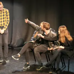 Młodzi aktorzy ćwiczą scenę. Jeden z nich wskazuje coś na widowni, jakby to był teatr na bruku.