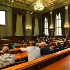 Sala obrad z ludźmi podnoszącymi ręce, widok zza pleców uczestników.