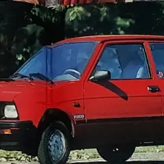 Czerwony yugo samochód z czarną atrapą chłodnicy i przyciemnianymi szybami, zaparkowany na tle zieleni.