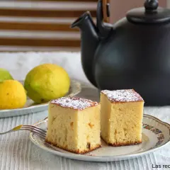 Dos porciones de bizcocho de limón esponjoso espolvoreadas con azúcar glas, junto a limones frescos y una tetera negra.