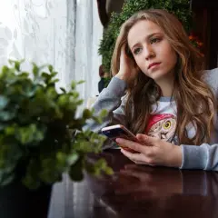 Zamyślona nastolatka z telefonem w dłoni, oparta o rękę, siedzi przy stoliku w kawiarni.
