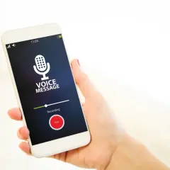 Dłoń trzyma smartfon z włączonym dyktafonem w telefonie. Na ekranie widnieje ikona mikrofonu i napis "VOICE MESSAGE".
