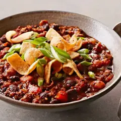 En skål med mustig, lakto-ovo vegetarisk chili med bönor, toppad med krispiga tortillachips och salladslök. En sked ligger bredvid.