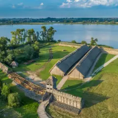 Rekonstrukcja osady Biskupin z drewnianymi domami krytymi strzechą, wałami obronnymi i jeziorem w tle.