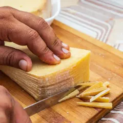 Mano cortando un bloque de queso en finas láminas. La corteza dorada se ve apetitosa, demostrando que se puede comer la corteza del queso.