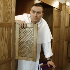 Młody zakonnik w białej sutannie otwiera kratkę konfesjonału, gotów wysłuchać spowiedzi.