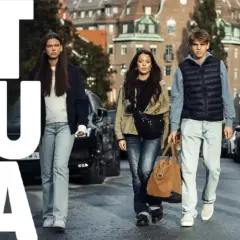 En grupp ungdomar går längs en gata. Stora bokstäver "STRULA" syns till vänster.
