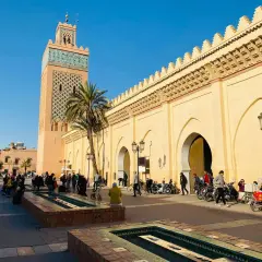 La moschea Koutoubia domina la piazza, con la sua torre decorata e le arcate del souk di Marrakech. Gente e motorini animano la scena.