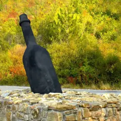 Giant black wine bottle sculpture emerging from a stone wall, with a colorful autumn forest in the background.