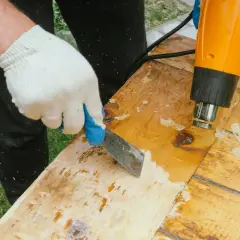 Main tenant un décapeur thermique et une spatule pour retirer la colle sur bois vernis.