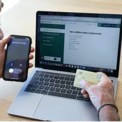 Un homme tient un téléphone et une carte bancaire, devant un ordinateur affichant le site du Crédit Agricole. Il cherche peut-on annuler un paiement par carte bancaire sur internet.