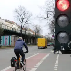 Ein Radfahrer wartet an einer roten Ampel. Im Hintergrund eine gelbe LKW und eine Hochbahn.