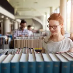 Studentka z rudymi włosami w okularach przegląda książki w bibliotece, szukając materiałów do prawo studia.