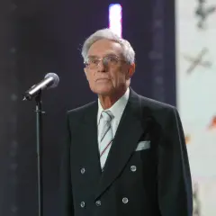 Starszy pan w okularach, Lucjan Szołajski, w czarnym garniturze i pasiastym krawacie stoi przy mikrofonie na tle sceny.