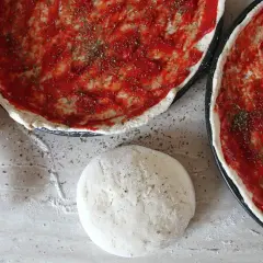Dwa surowe ciasta na pizzę z sosem pomidorowym i ziołami, wałek i kulka ciasta.