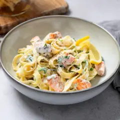 Pyszny makaron z łososiem w kremowym sosie, udekorowany cytryną i natką pietruszki. Obok leżą surowe tagliatelle.