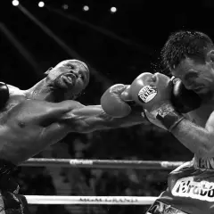 A boxer throws a powerful double jab, sweat flying as his opponent braces for impact in the intense spotlight of the ring.