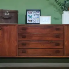 Mobile antico in legno scuro, con cassetti e ante. Sopra, una valigia vintage, quadri di farfalle e un vaso con fiori. Guida su come pulire un mobile antico.
