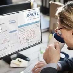 Mężczyzna pije kawę, patrząc na ekran komputera z fakturą z pieczątką "PAID" i informacją o podatku VAT.